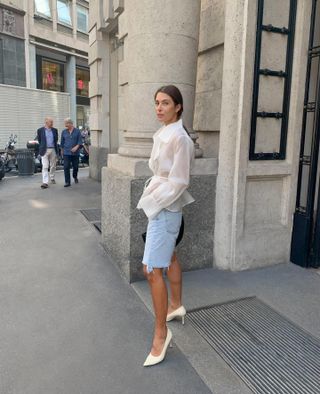 Woman wears long denim shorts, organza top, and white kitten heels.