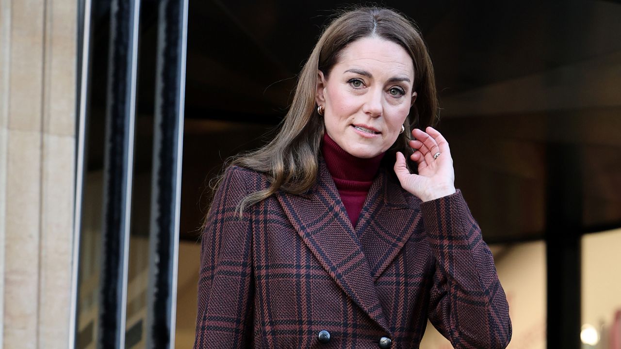 Kate Middleton wearing a plaid coat and red turtleneck lifting one hand up to her face