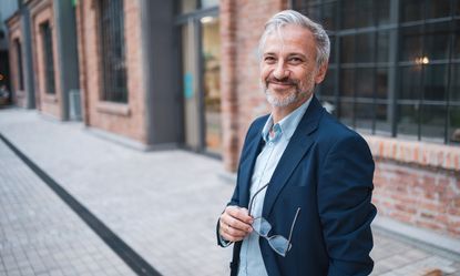 A mature businessman stands confidently outdoors in an urban environment, wearing a suit and holding his glasses. His warm smile conveys positivity and professional success. Stylish brick buildings are visible in the background.