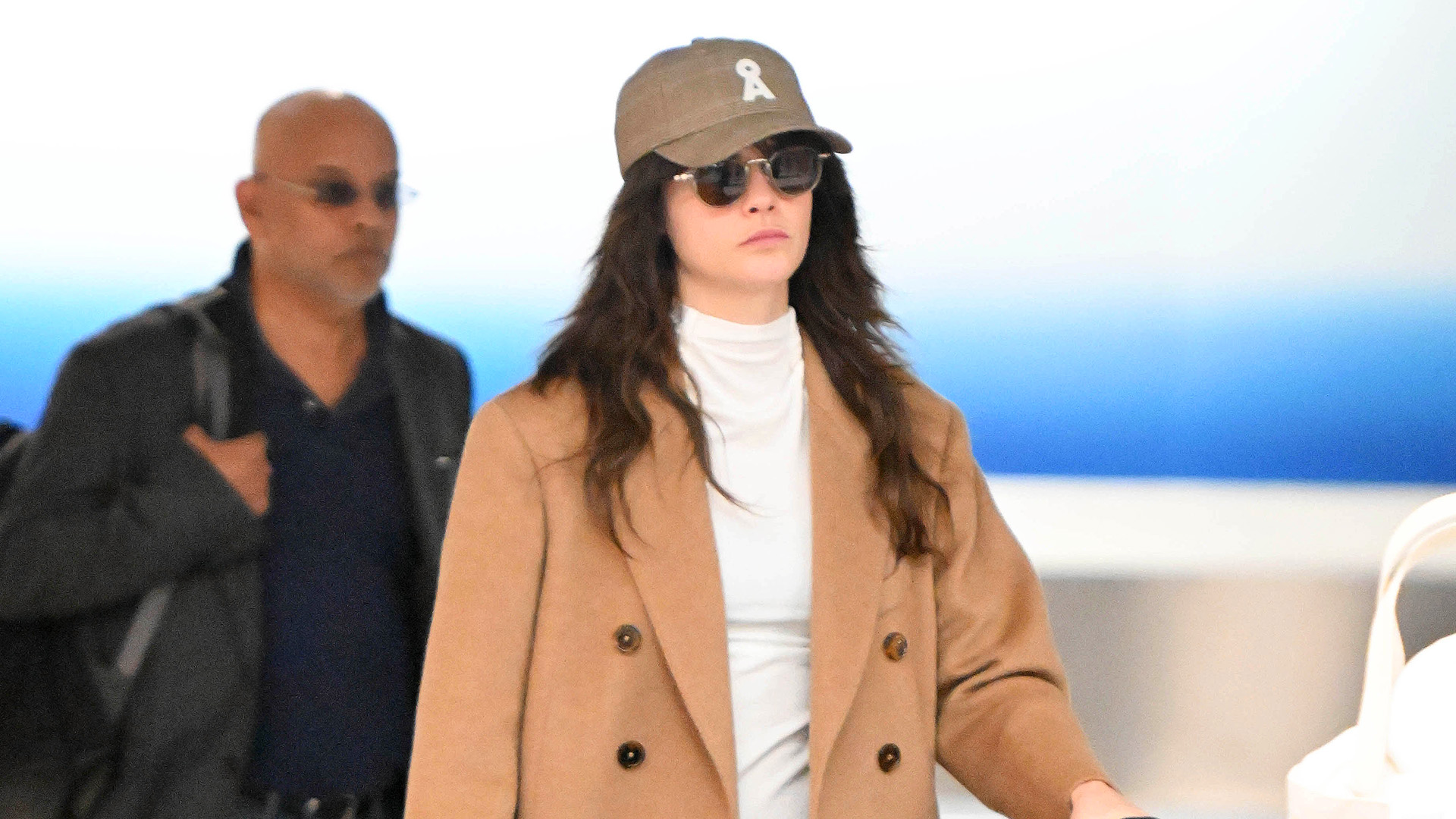 Cara Delevingne wearing cobalt blue pants with a camel coat, cream turtleneck and black suede mules at JFK airport
