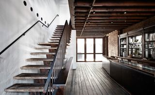 Long room, wooden floor, wooden stairwell with black hand rail, white wall to the left, brick wall and bar area to the right, far end high wooden framed windows letting in light, dark wood ceiling