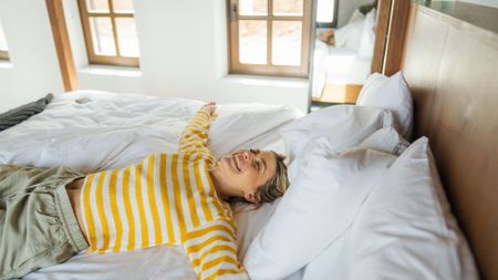 A woman wearing a yellow stripey top stretched out in a starfish position on a bed smiling