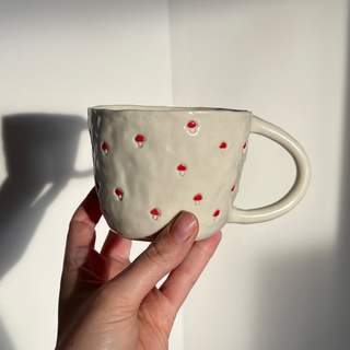 A hand holding a white coffee cup with dainty red mushroom pattern, in front of a plain white background.