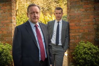 Midsomer murders - DCI John Barnaby (Neil Dudgeon) and DS Jamie Winter (Nick Hendrix) stand in their work suits under a red brick archway in a country house garden