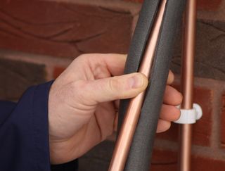 A mans hand wrapping a copper pipe in a coat of foam insulation against a brick wall