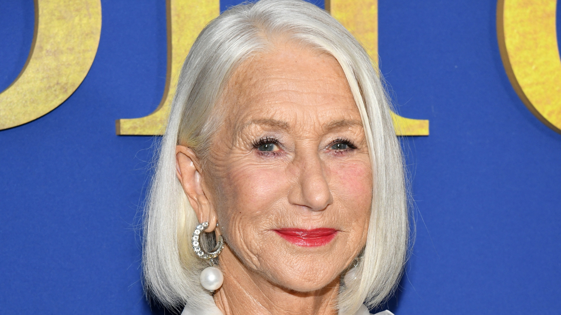 Helen Mirren smiling with a side part grey bob and wearing fuchsia lipstick, on a blue background
