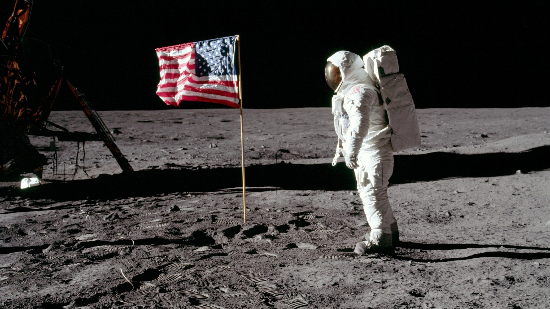 an astronaut in a white space suit looks at a flag planted on a dusty grey surface