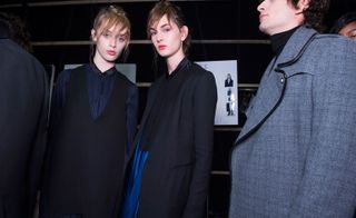 Two female models wearing looks from Costume National's collection. One model is wearing a dark blue top and dark blue V-neck sleeveless piece. Next to her is a model wearing a black and blue piece with a dark blue jacket over the top. And there is a third male model wearing a black turtle neck and grey jacket with black trims