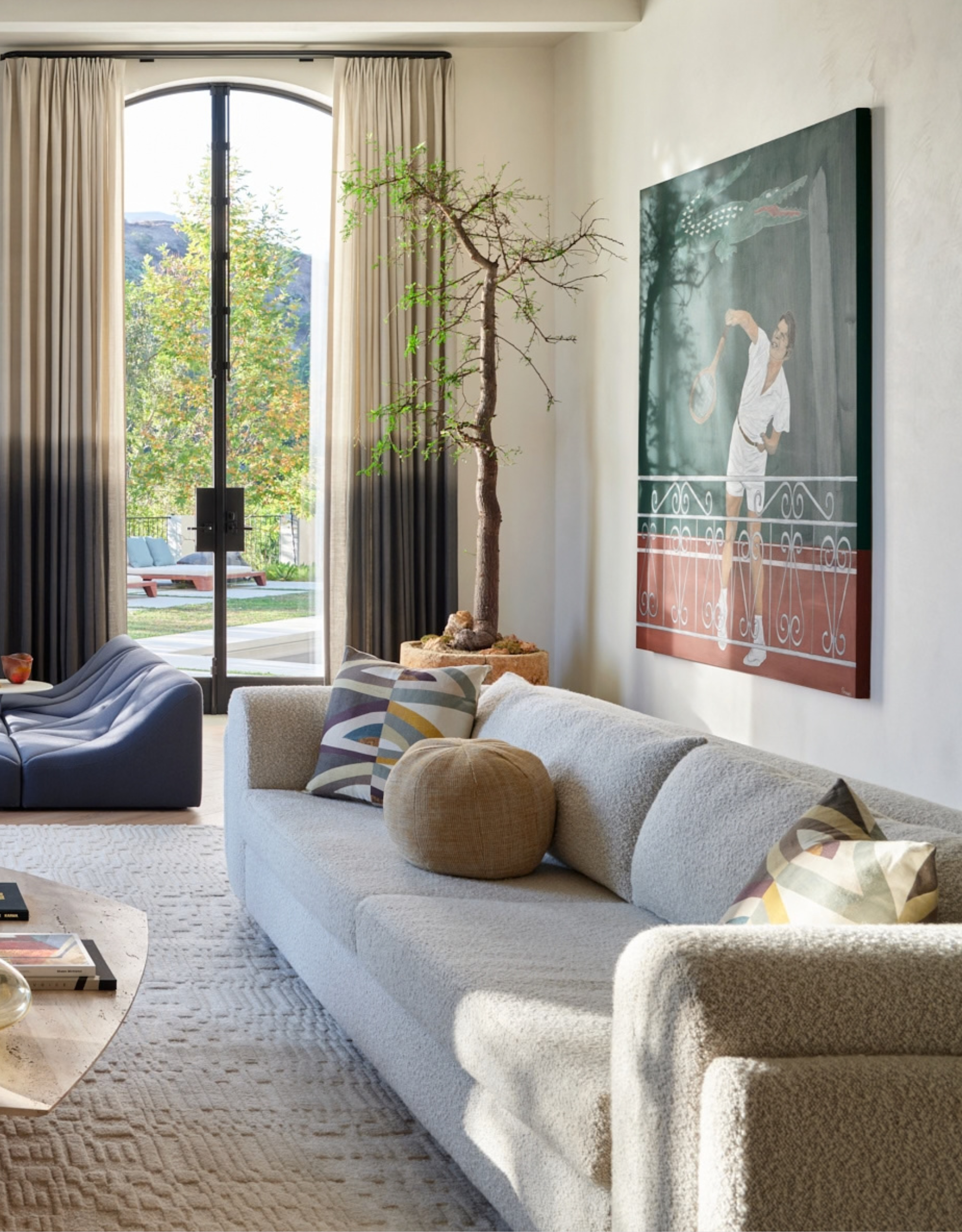 A living room with two-toned curtains, a potted tree, a boucle sofa, and a tennis-themed painting
