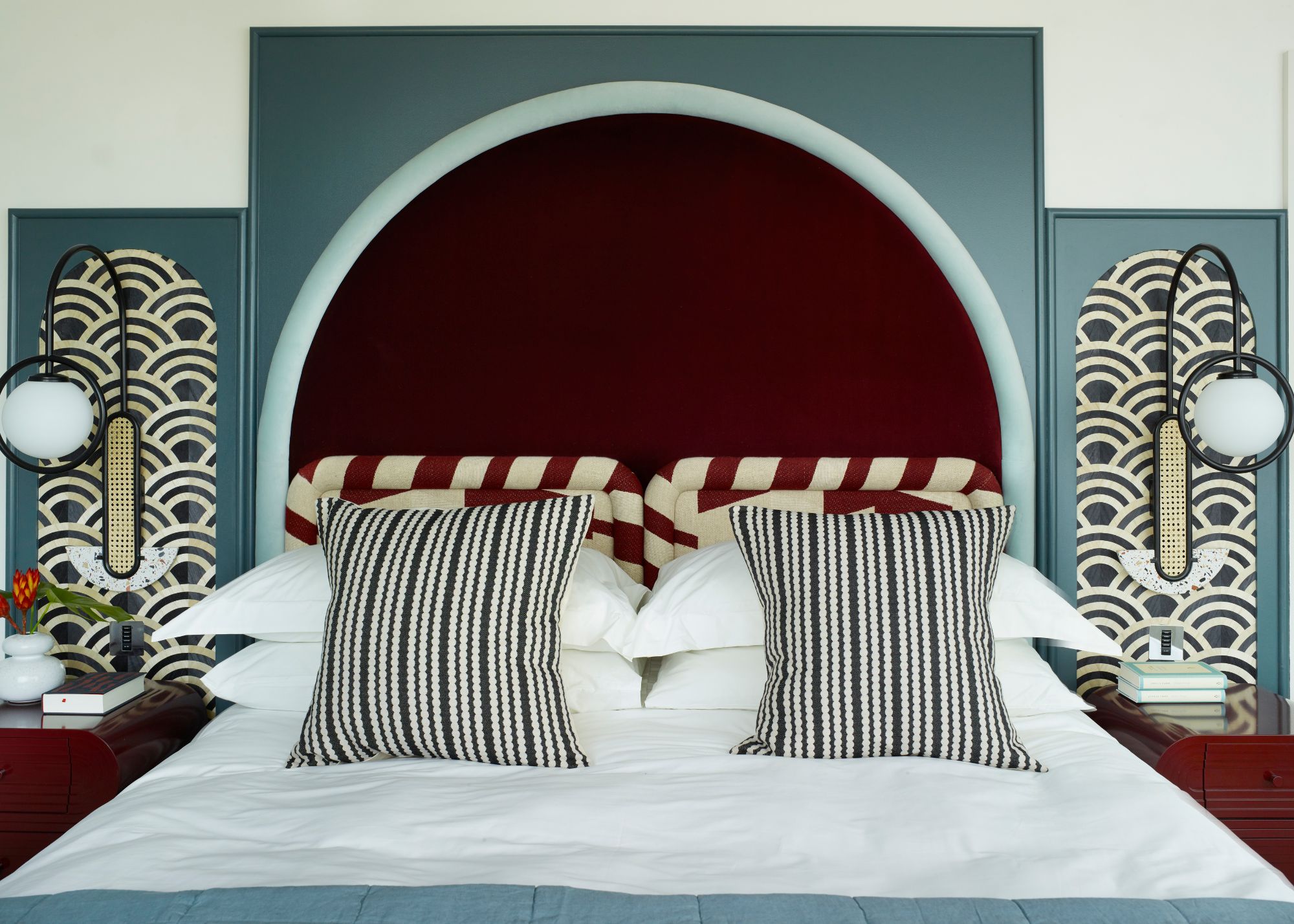 A double bed with a statement dark red arched headboard, set on blue wall panelling. Two further arches above each nightstand with geometric wallpaper.