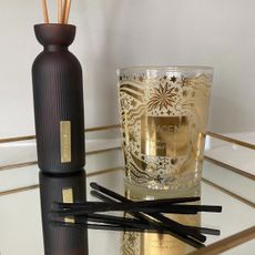 Close up of the Space NK Shimmering Spice Candle sat on a gold edged mirror tray, a black reed diffuser to the left and black matches at the front 