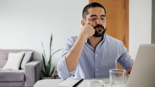 A short-bearded man in a business shirt sitting at a desk in front of a laptop, leaning on his elbow and looking bored.