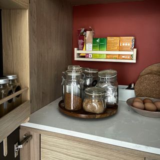 Image of the inside of a larder unit with white worktop, orange wall and wooden lazy susan holding glass jars