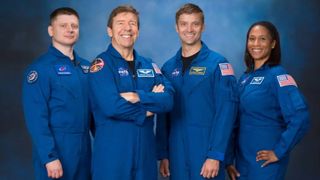 four smiling astronauts in blue flight suits stand in front of a blue background