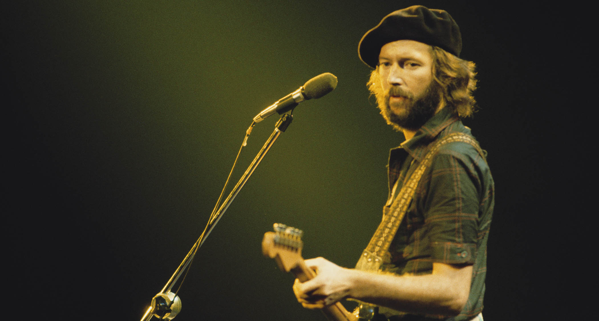 Eric Clapton wears a beret and plays a Fender Stratocaster onstage.