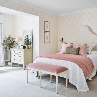 a bedroom with a bed with a large cream upholstered headboard, a pink velvet upholstered footstool at the end of the bed with a matching throw on the bed and a cream chest of drawers with mini Christmas tree