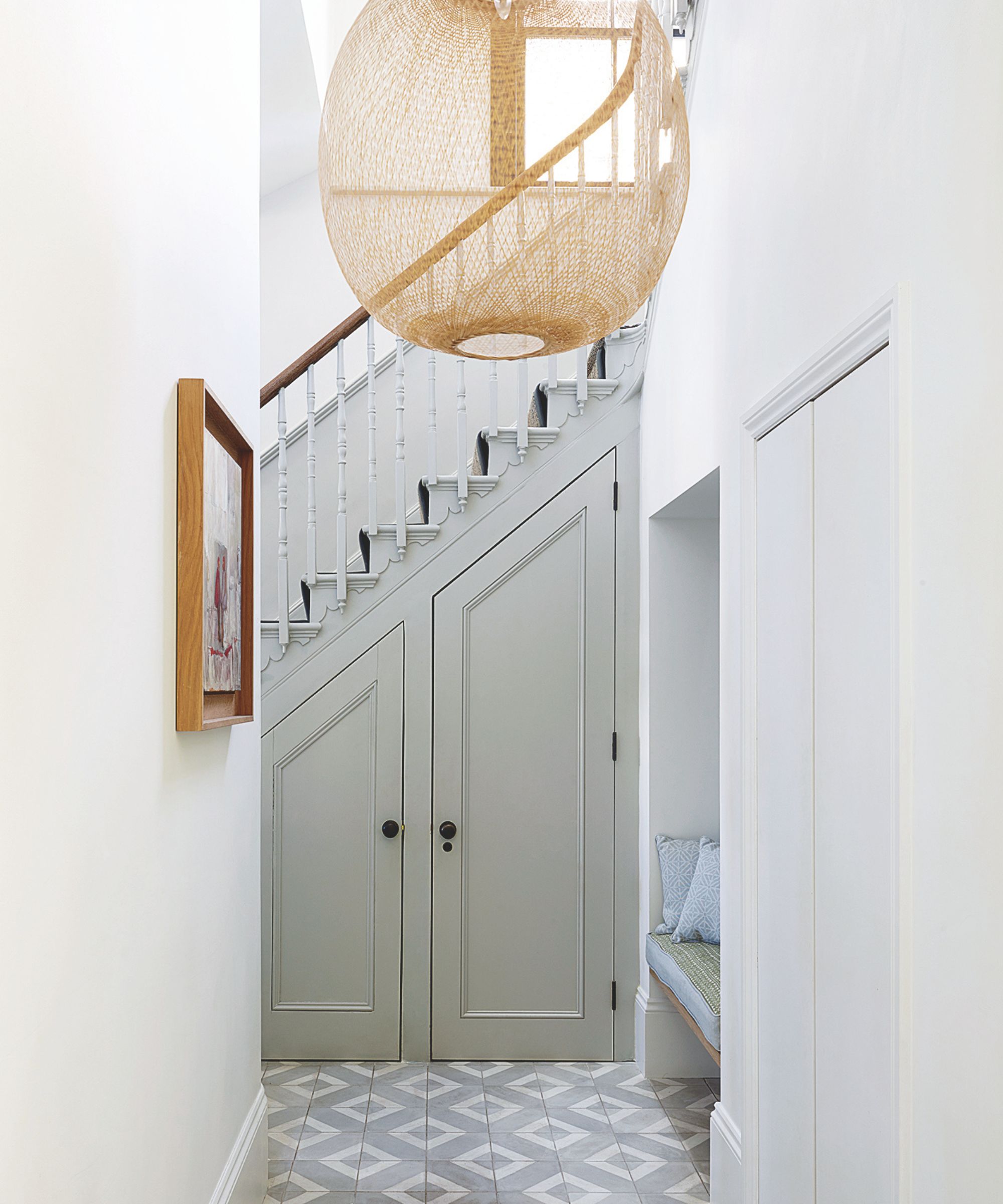 A white entryway with tiled floors, a rattan lamp, in-built bench seating, a wooden framed artwork on the wall, and a large rattan lamp hanging above.
