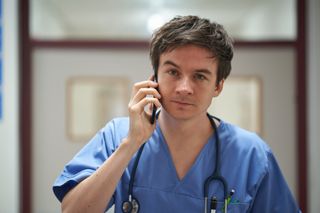 Scott Chambers as Dr Oscar Beattie on the phone wearing scrubs and a stethoscope