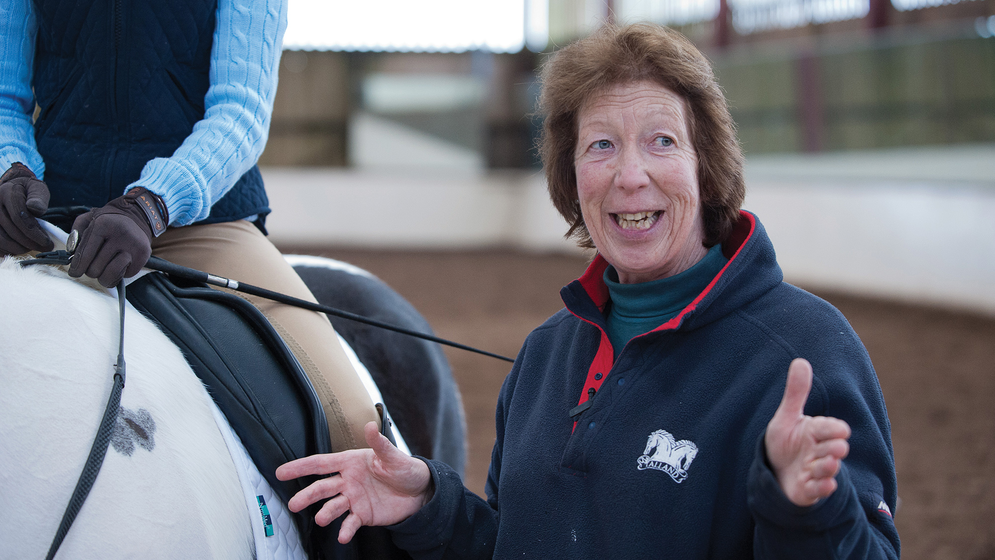 Pammy Hutton stands beside a young rider on a coloured horse with her hands open explaining a lesson. 