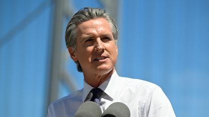 A telephoto shot of Gavin Newsom, governor of California, stood against the backdrop of the San Francisco&ndash;Oakland Bay Bridge with a bright blue sky visible in the background.