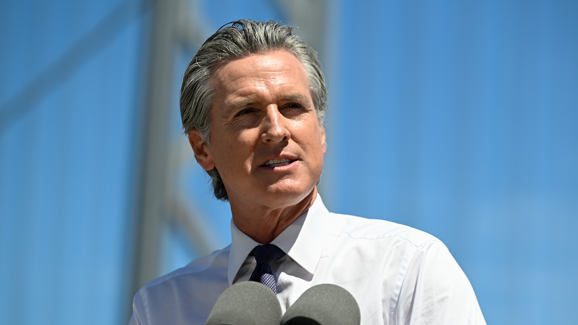 A telephoto shot of Gavin Newsom, governor of California, stood against the backdrop of the San Francisco–Oakland Bay Bridge with a bright blue sky visible in the background.