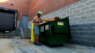 A shirtless man in tan cargo shorts with a neon yellow bike helmet pulls bananas out of a green dumpster.
