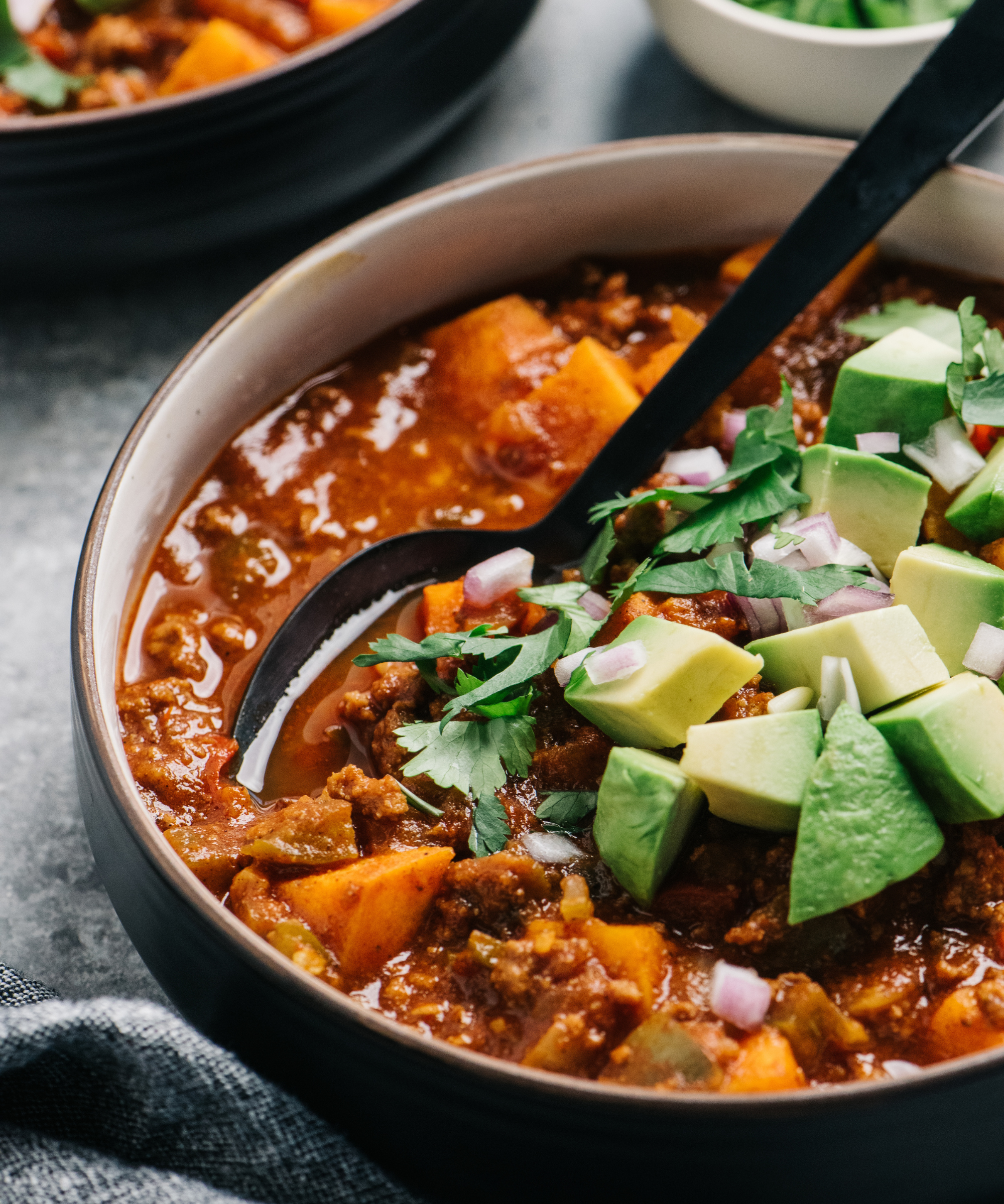 A bowl of chilli con carne with chunky vegetables, topped with diced avocado and cilantro