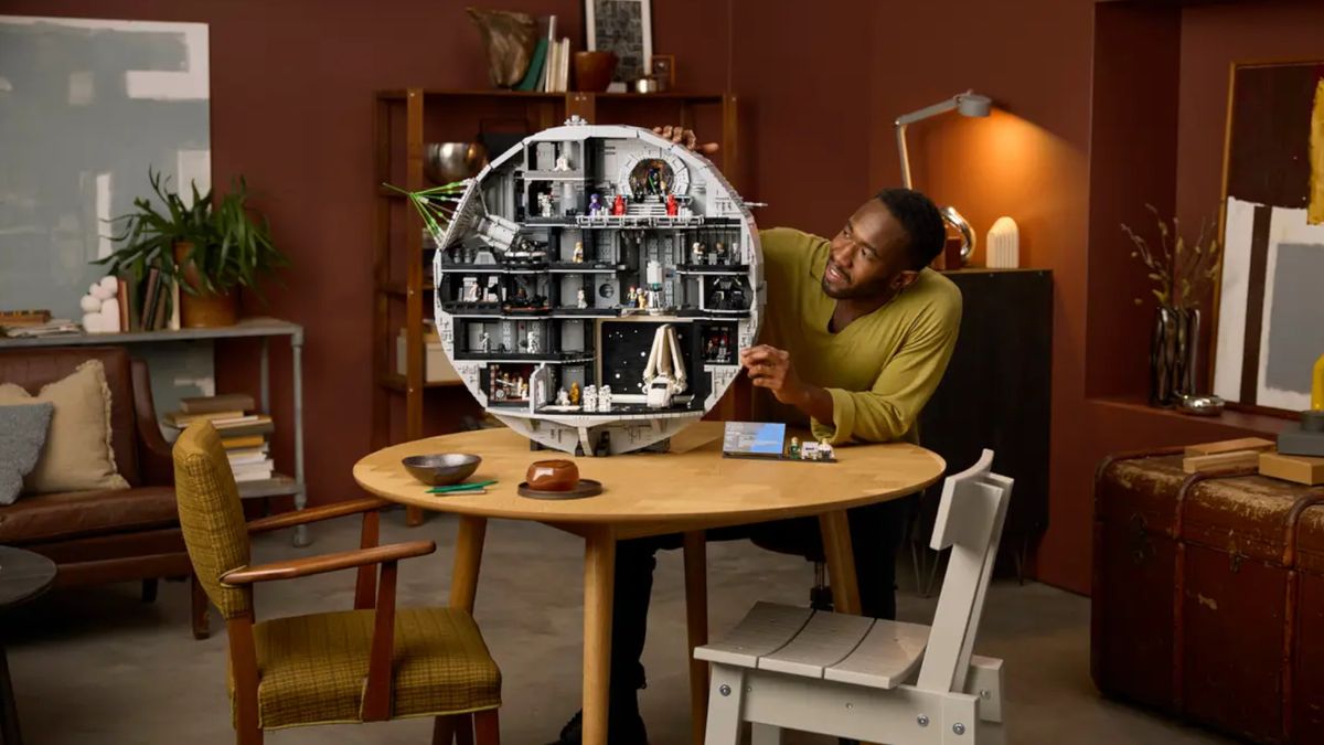 A smiling man places pieces on the Lego Death Star model while sitting at a wooden table