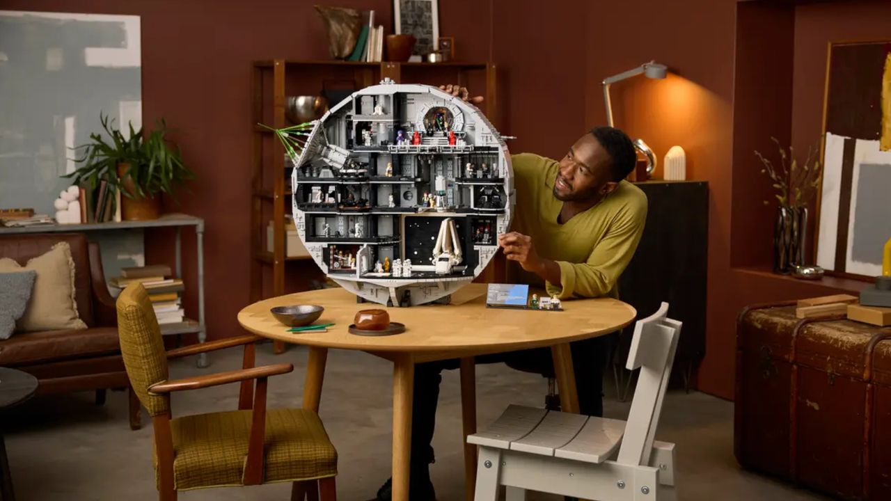 A smiling man places pieces on the Lego Death Star model while sitting at a wooden table
