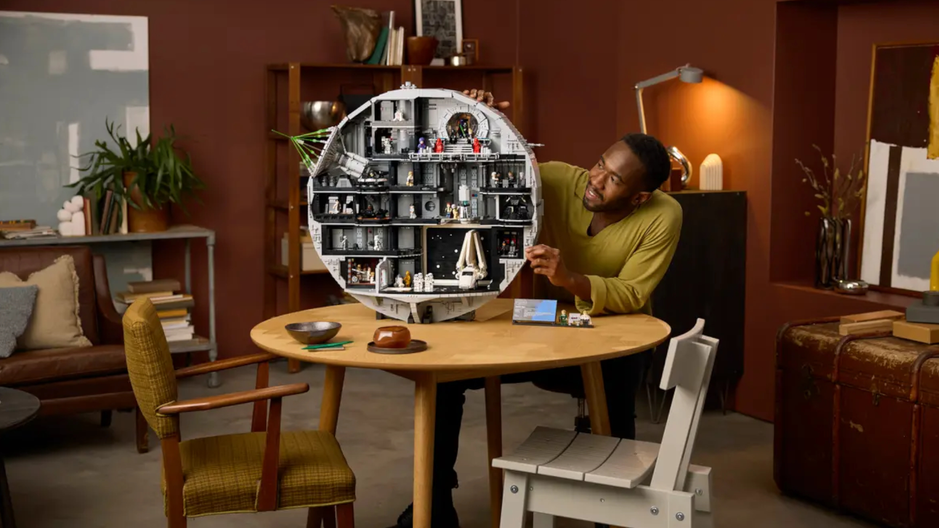 A smiling man places pieces on the Lego Death Star model while sitting at a wooden table