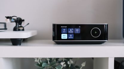 A lifestyle photo of the WiiM Amp Ultra on top of a white unit with a white and black turntable beside it