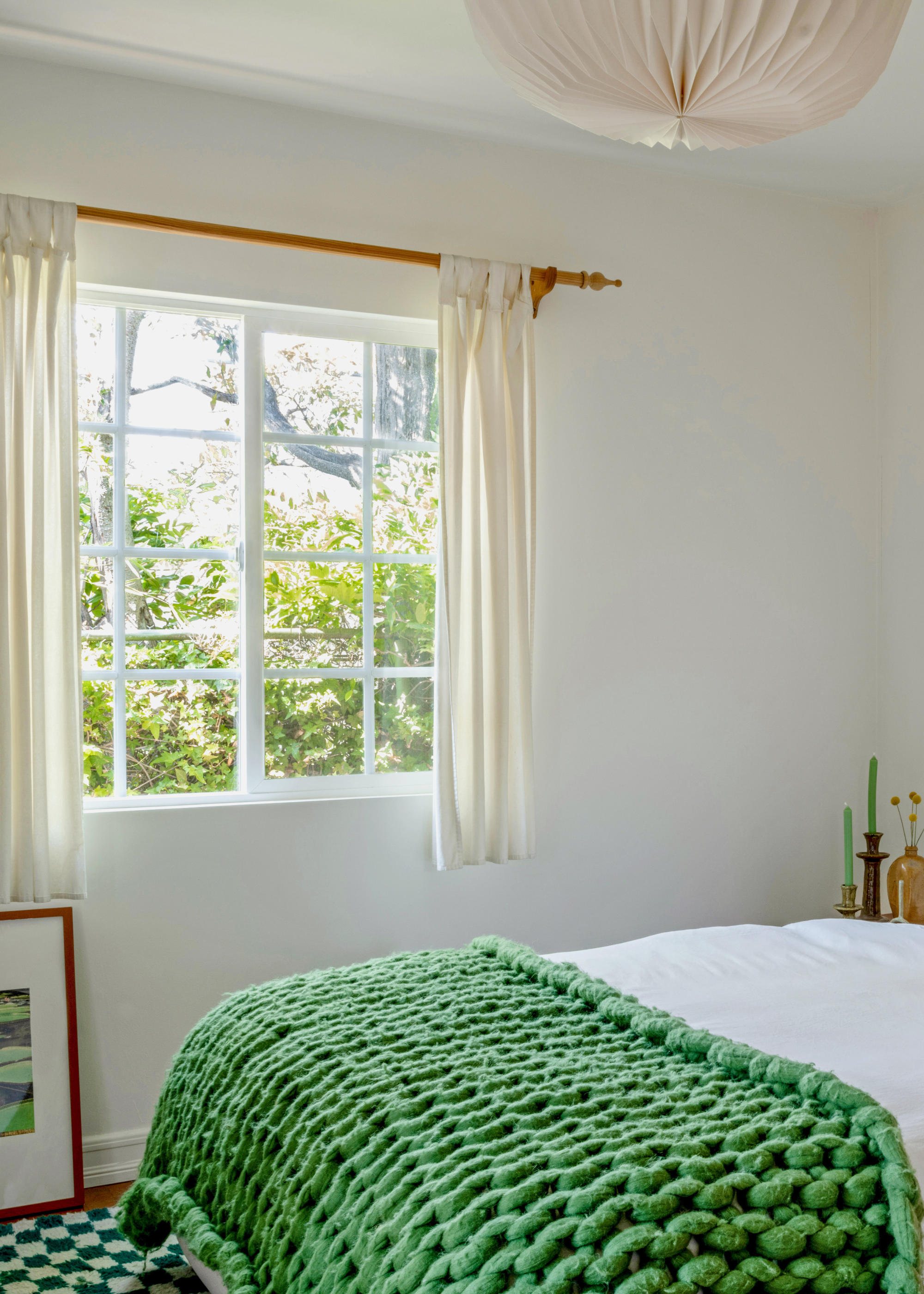 A white bedroom with large windows and sheer curtains, a framed painting on the floor, a white bed with a thick woven green knit throw blanket and a green checkerboard rug