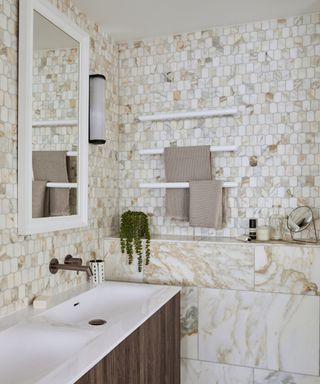 A bathroom with white and gold marbled walls, a white wooden framed mirror, a white sink with dark brown wooden cabinets, and three towel bars with brown towels folded on them, and a mirror, plant, and toiletries
