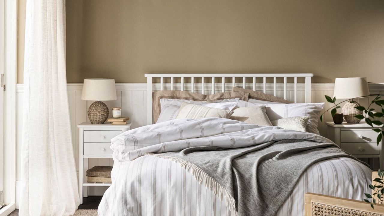 A warm toned bedroom with half panelled walls. A white bed frame, grey bedding. Matching white bedside table with a small stone lamp. 