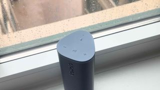 The Sonos Roam 2 stands upright on one of its ends, by a window. We can see the controls on the top of the device, which control playback and the voice assistant feature.