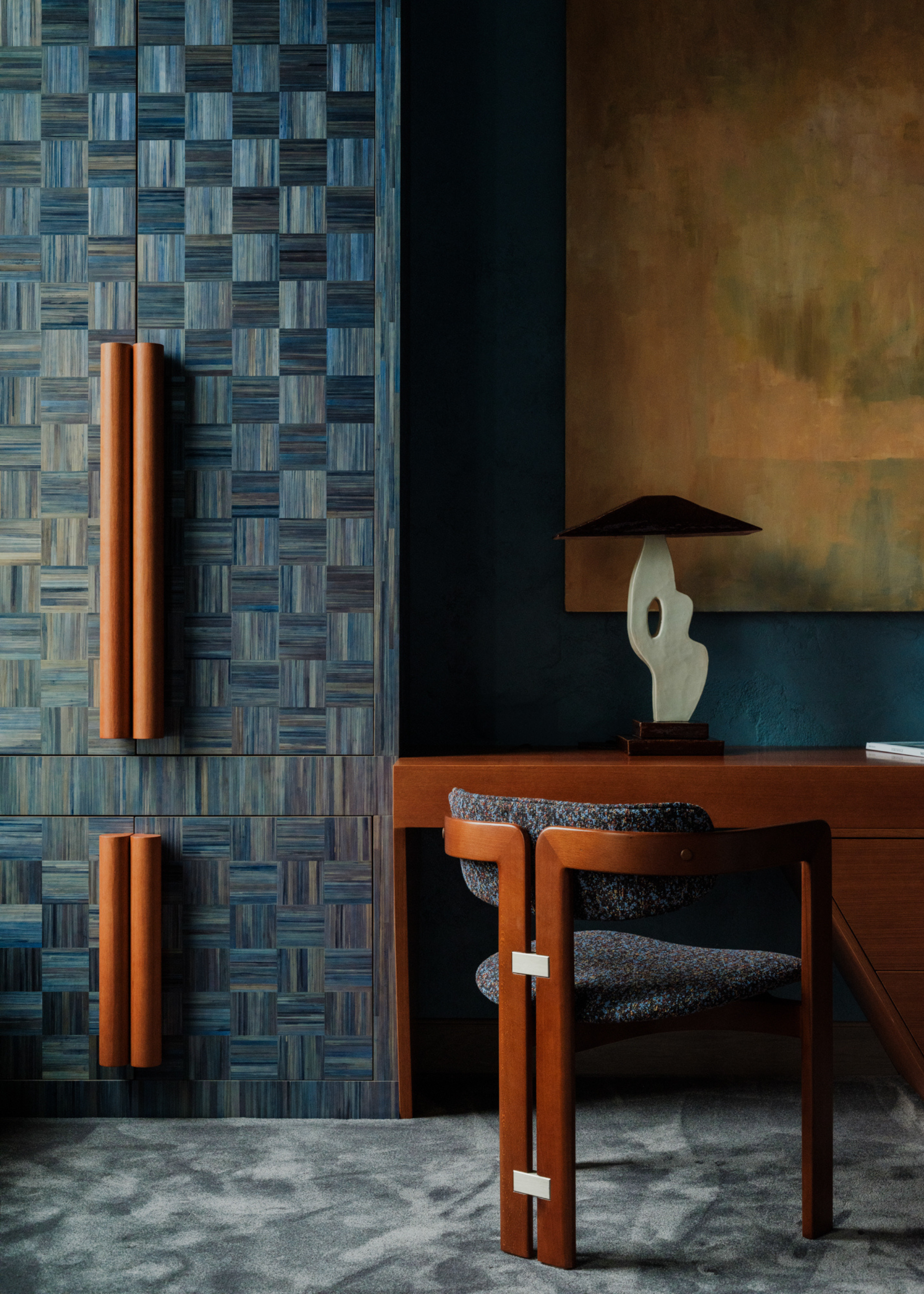 A bedroom with blue wood cupboards by a wood study with an armchair with a desk lamp and a framed painting