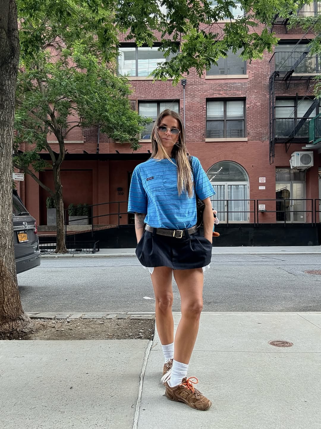 Janka Polliani is wearing a black mini skirt. She pairs it with a blue striped t-shirt, white sock and the Miu Miu X New Balance collab trainer. She is stood posing into the camera in this image taken from her Instagram recently, and is stood outside some red brick buildings in New York.