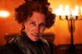 A close up of Tamsin Greig in character as Mrs Wickens, wearing a curly red wig and with her face made-up to look red and blotchy