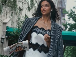 model wearls white slip dress with lace details and a long gray trench coat while holding a newspaper and walking