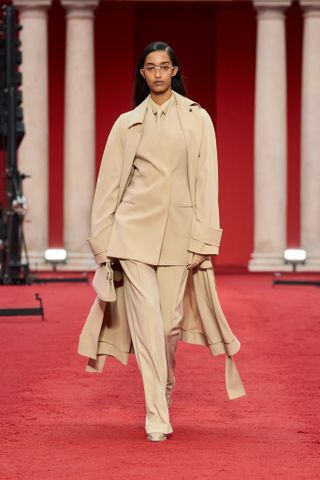 A female model wearing a light brown suite and coat walking on a red carpet.