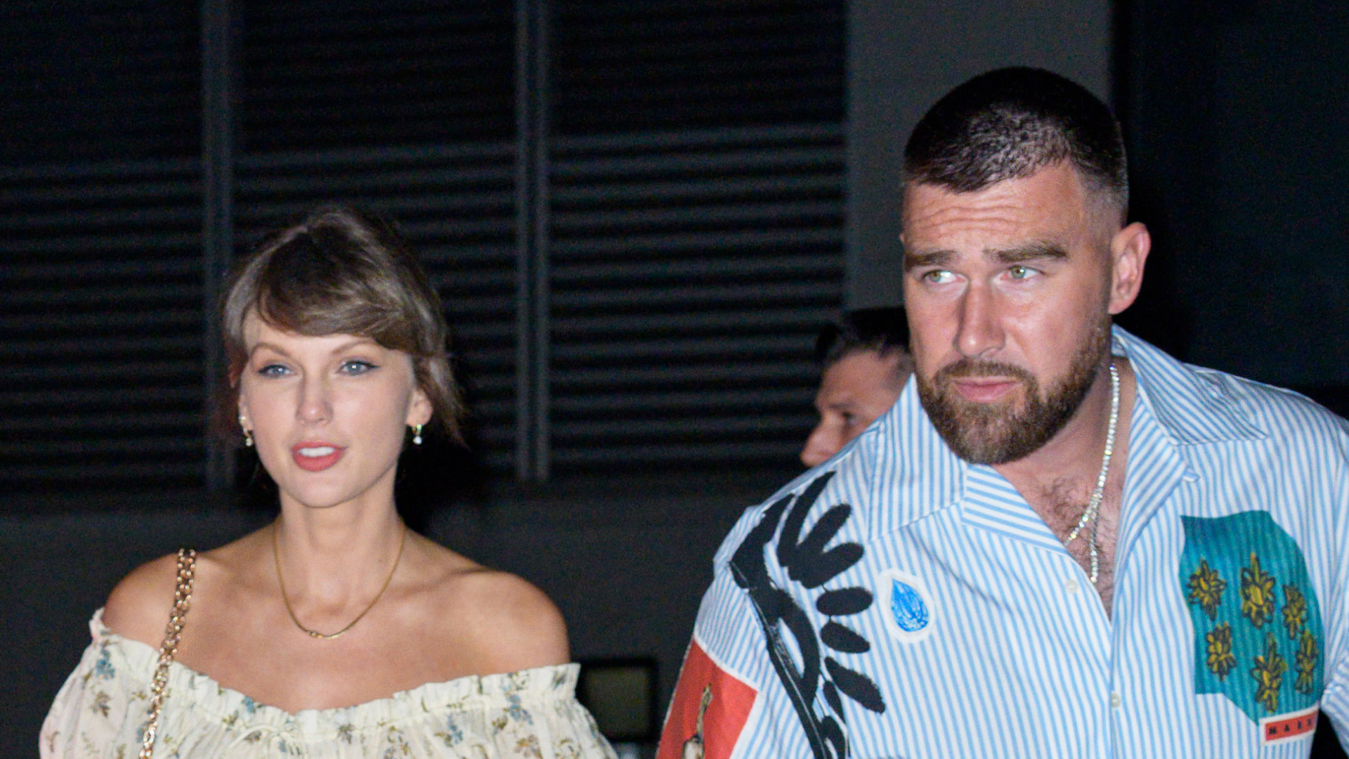 Taylor Swift wears a floral off-the-shoulder dress and holds hands with her husband Travis Kelce who wears a striped shirt