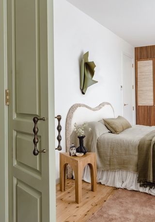 Photo entering a white and sage green bedroom. The bedding is a linen material with hardwood floors and a wooden side table also in the room