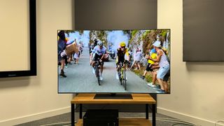 The 65-inch LG G4 OLED TV on a wooden TV stand. On screen are two cyclists with spectators cheering them on from the side of the road.