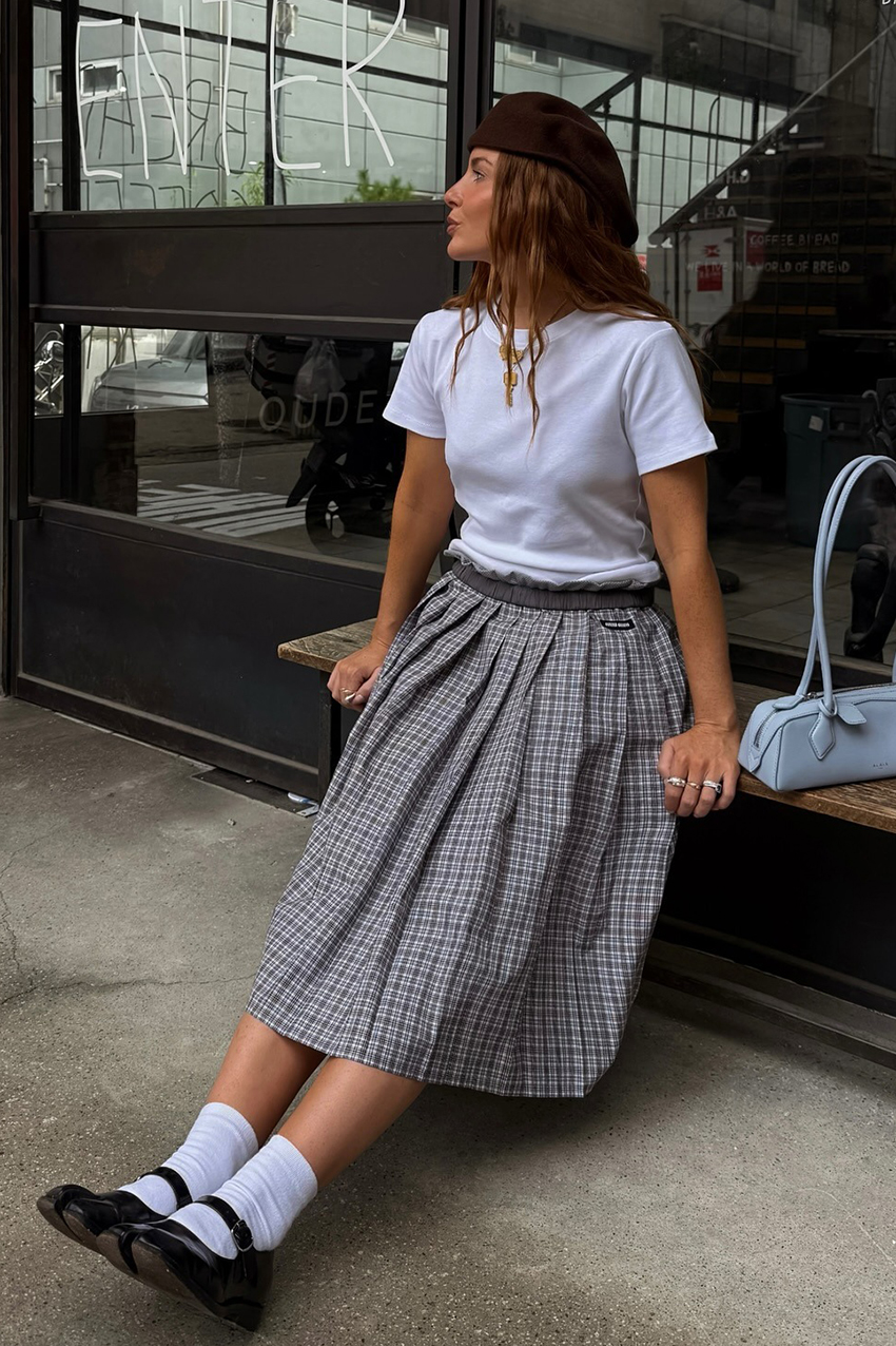 a woman wearing a white t-shirt, plaid skirt, white socks, black mary janes, and brown flat cap