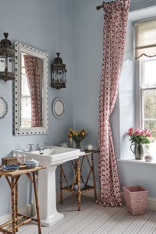 An example of blue bathroom ideas showing a pastel blue bathroom in period setting, pink print curtains, an ornate mirror, wall lights, and rattan side units