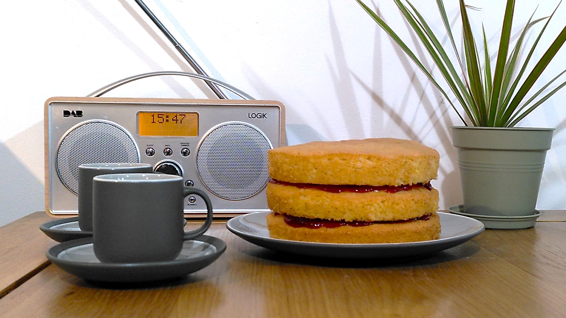 Three-layer Victoria sponge cake cooked using air fryer on table beside radio, potted plant, and two coffee cups