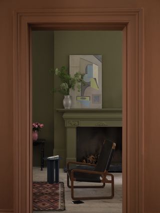 A sage green living room as seen from the doorway that is painted in a dark orange color. There is a fireplace in the living room with a gray vase full of green flowers and a gray, blue, and green abstract painting. There is a black leather and wooden accent chair in front of the fireplace