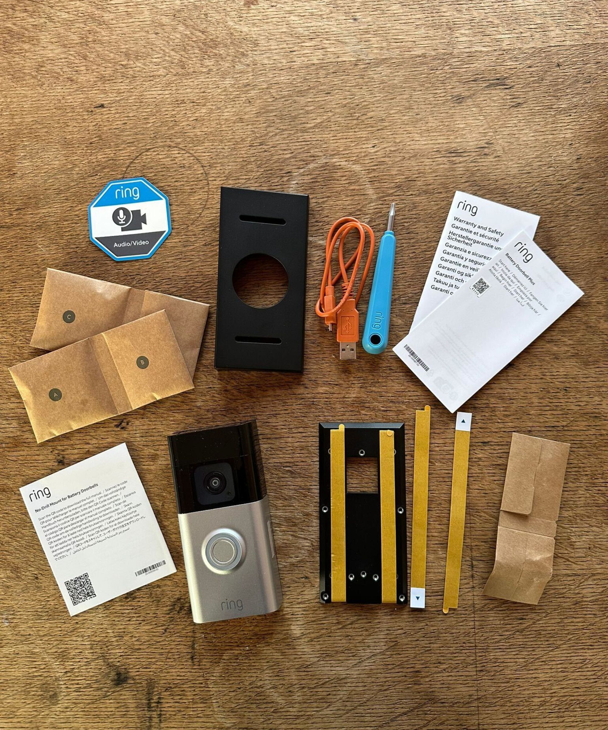 The contents of a Ring Battery Doorbell Plus box, including the doorbell, envelopes of screws, paper instructions and blue screw, unboxed and laid out on a wooden surface.