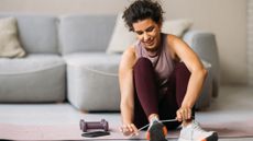 A woman sits on a yoga mat in her living room tying up her shoes, next to her is a pair of light dumbbells and a phone