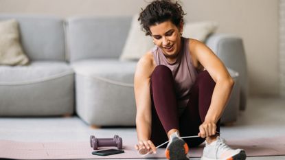 A woman sits on a yoga mat in her living room tying up her shoes, next to her is a pair of light dumbbells and a phone